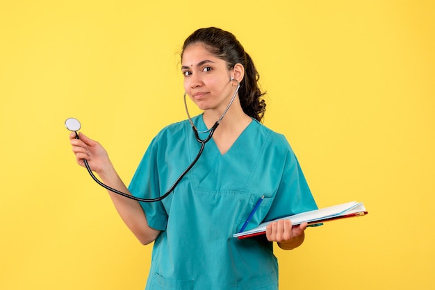 Vooraanzicht jonge vrouwelijke arts in eenvormige holdingsstethoscoop en klembord op gele achtergrond