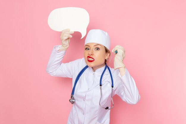Vooraanzicht jonge vrouwelijke arts in wit medisch pak met blauwe stethoscoop injectie en wit bord op de roze ruimte geneeskunde medische ziekenhuis werknemer te houden