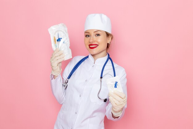 Vooraanzicht jonge vrouwelijke arts in wit medisch pak met blauwe stethoscoop injectie pakket houden op het roze ruimte geneeskunde medische ziekenhuis