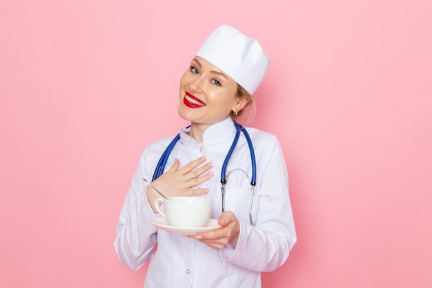 Vooraanzicht jonge vrouwelijke arts in wit medisch pak met blauwe stethoscoop kopje koffie met glimlach op de roze ruimte geneeskunde medische ziekenhuis te houden