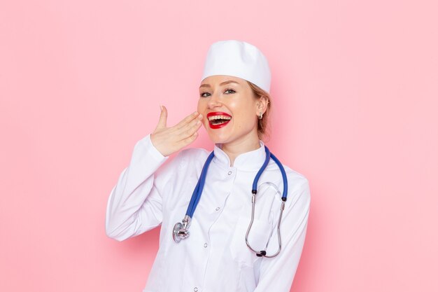 Vooraanzicht jonge vrouwelijke arts in wit pak met blauwe stethoscoop poseren en glimlachend op de roze ruimtebaan