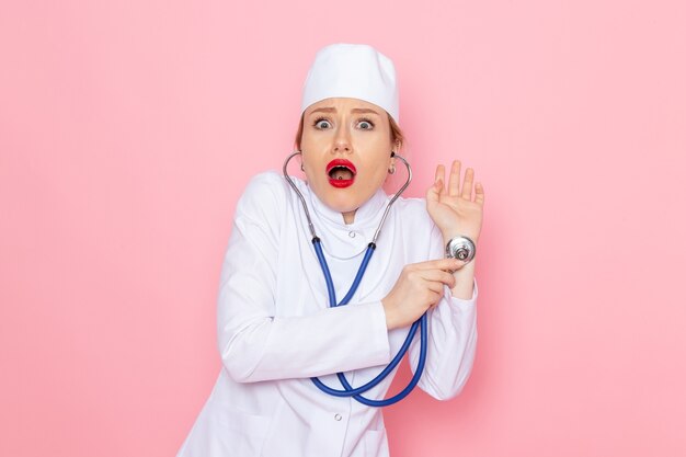 Vooraanzicht jonge vrouwelijke arts in wit pak met blauwe stethoscoop poseren meten met verbaasde uitdrukking op de roze ruimte geneeskunde medische ziekenhuis