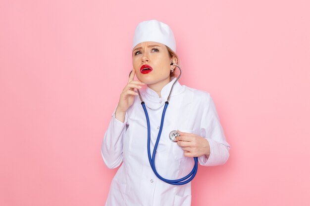 Vooraanzicht jonge vrouwelijke arts in wit pak met blauwe stethoscoop poseren meten op de roze ruimte geneeskunde medisch ziekenhuis vrouwelijk werk