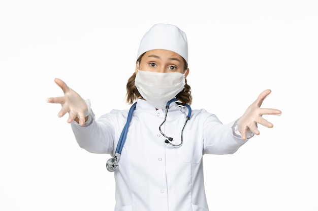 Vooraanzicht jonge vrouwelijke arts met steriel masker en handschoenen vanwege coronavirus op wit oppervlak