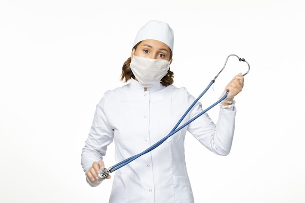 Vooraanzicht jonge vrouwelijke arts met steriel masker vanwege coronavirus op het lichtwitte oppervlak