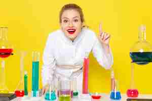 Gratis foto vooraanzicht jonge vrouwelijke chemicus in wit pak voor tafel met ed-oplossingen glimlachend op het gele ruimtechemie wetenschappelijk experiment