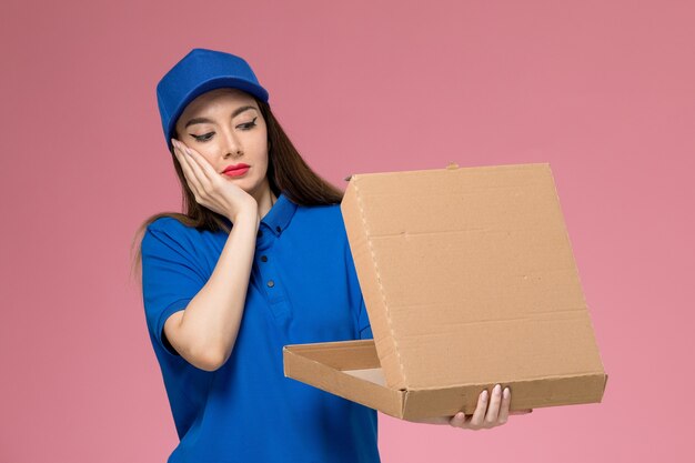 Vooraanzicht jonge vrouwelijke koerier in blauw uniform en cape met voedseldoos met verwarde uitdrukking op de roze muur