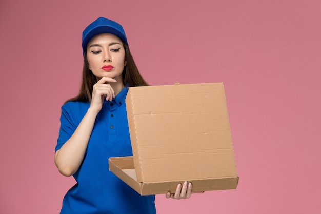 Vooraanzicht jonge vrouwelijke koerier in blauw uniform en cape met voedselleveringsdoos denken aan de roze muur