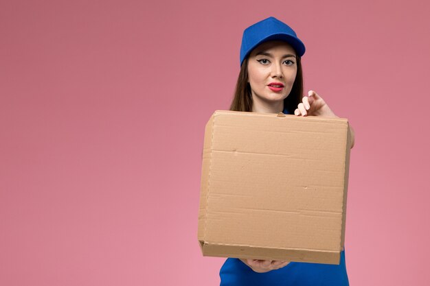 Vooraanzicht jonge vrouwelijke koerier in blauw uniform en cape met voedselleveringsdoos op de lichtroze muur