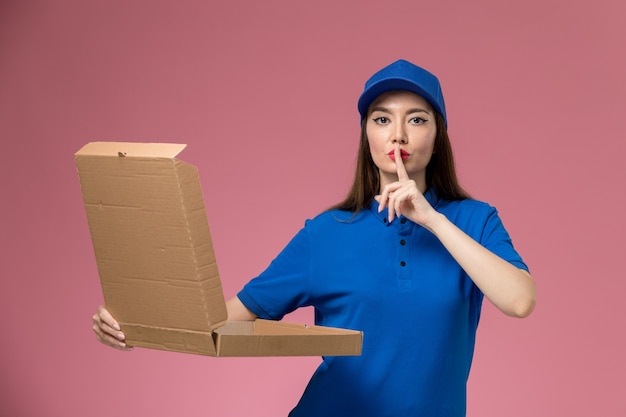 Vooraanzicht jonge vrouwelijke koerier in blauw uniform en cape met voedselleveringsdoos op lichtroze muurdienst dame levering uniform baan