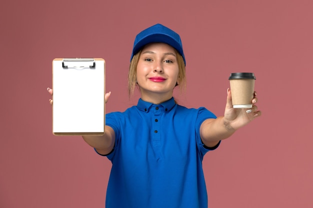 Vooraanzicht jonge vrouwelijke koerier in blauw uniform poseren bedrijf kopje koffie en blocnote glimlachen, service uniforme levering vrouw baan werknemer kleur