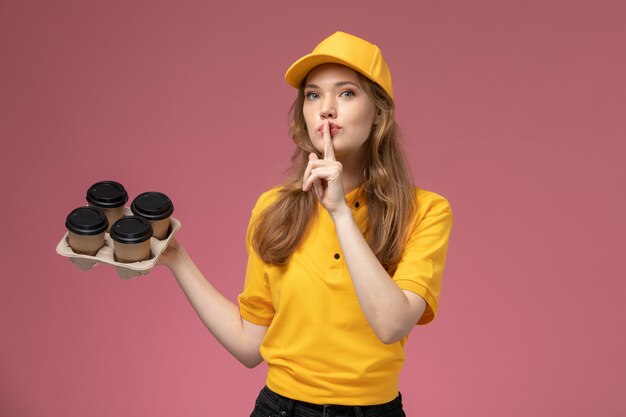 Vooraanzicht jonge vrouwelijke koerier in geel uniform bedrijf koffiekopjes stilte teken op donker roze bureau baan uniforme levering service werknemer tonen