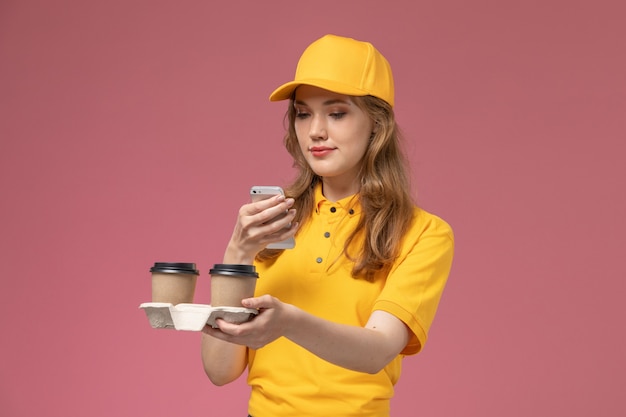 Vooraanzicht jonge vrouwelijke koerier in geel uniform bezorgen koffie houden en met behulp van haar telefoon op de roze bureau baan uniforme bezorgdienst werknemer