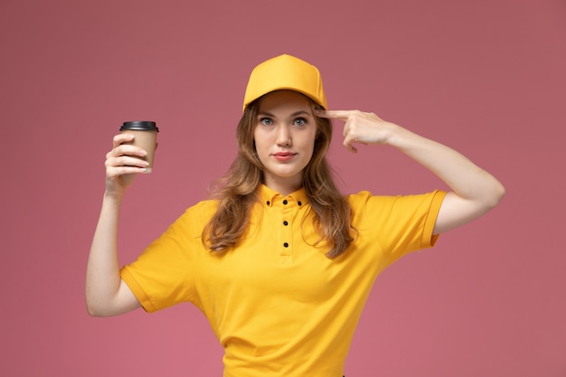 Vooraanzicht jonge vrouwelijke koerier in geel uniform bezorgen koffie houden en poseren op de roze achtergrond baan uniforme bezorgdienst werknemer