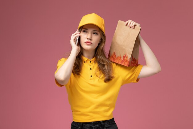 Vooraanzicht jonge vrouwelijke koerier in geel uniform levering voedselpakket houden en praten aan de telefoon op roze bureau baan uniforme bezorgdienst werknemer