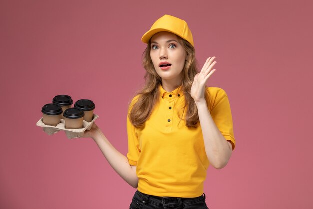 Vooraanzicht jonge vrouwelijke koerier in geel uniform met plastic bruine koffiekopjes op de roze-donkere achtergrond baan uniforme levering kleur dienstverlener