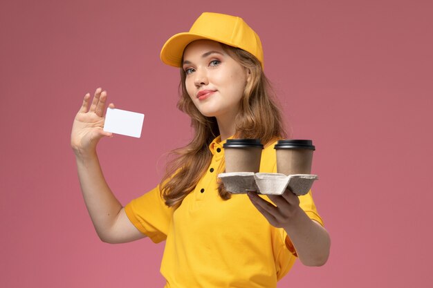 Vooraanzicht jonge vrouwelijke koerier in geel uniform met plastic koffiekopjes en witte kaart op donkerroze bureau uniforme bezorgdienst werknemer