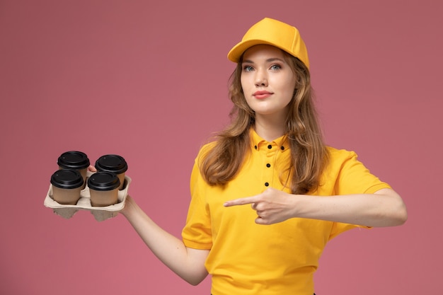 Vooraanzicht jonge vrouwelijke koerier in geel uniform met plastic koffiekopjes op de donkerroze achtergrond uniforme levering baan dienstverlener