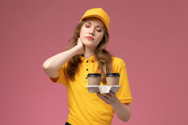 Vooraanzicht jonge vrouwelijke koerier in gele uniforme koffiekopjes met hoofdpijn op roze achtergrond bureau baan uniforme bezorgdienst werknemer