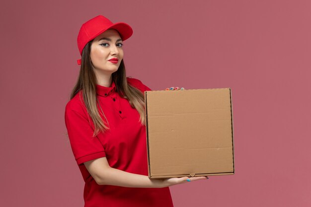 Vooraanzicht jonge vrouwelijke koerier in rode uniforme levering voedsel doos met glimlach op roze achtergrond service levering baan uniform bedrijf