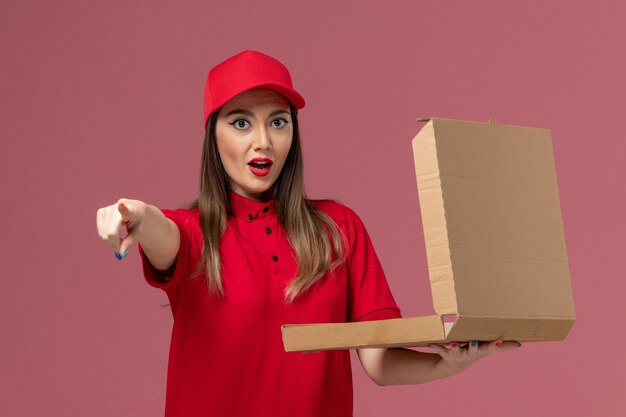 Vooraanzicht jonge vrouwelijke koerier in rode uniforme levering voedseldoos op het lichtroze bureau service levering baan uniform bedrijf