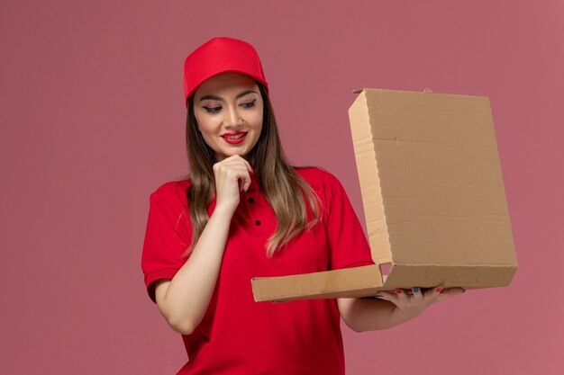 Vooraanzicht jonge vrouwelijke koerier in rood uniform bedrijf levering voedsel doos en denken over lichtroze achtergrond service levering uniform werknemer bedrijf