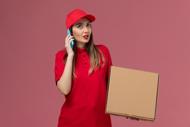 Vooraanzicht jonge vrouwelijke koerier in rood uniform bedrijf levering voedsel doos tijdens het praten aan de telefoon op roze achtergrond bezorgdienst uniform bedrijf