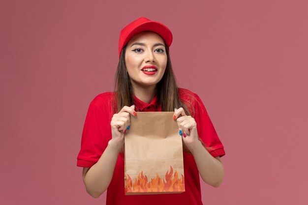 Vooraanzicht jonge vrouwelijke koerier in rood uniform bedrijf papier voedselpakket op roze achtergrond werknemer dienstverlening levering uniform bedrijf baan