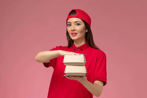 Vooraanzicht jonge vrouwelijke koerier in rood uniform en cape met kleine bezorgvoedselpakketten op haar handen op de roze muur