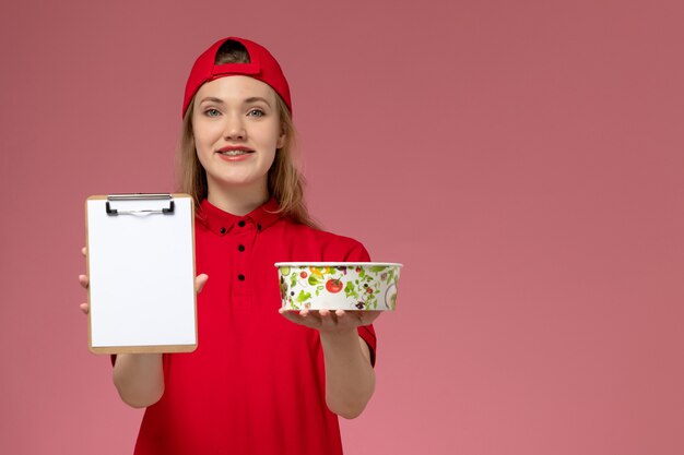 Vooraanzicht jonge vrouwelijke koerier in rood uniform en cape met leveringskom en blocnote met glimlach op roze muur