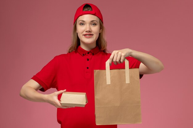 Vooraanzicht jonge vrouwelijke koerier in rood uniform en cape met voedselpakketten voor levering op de roze muur