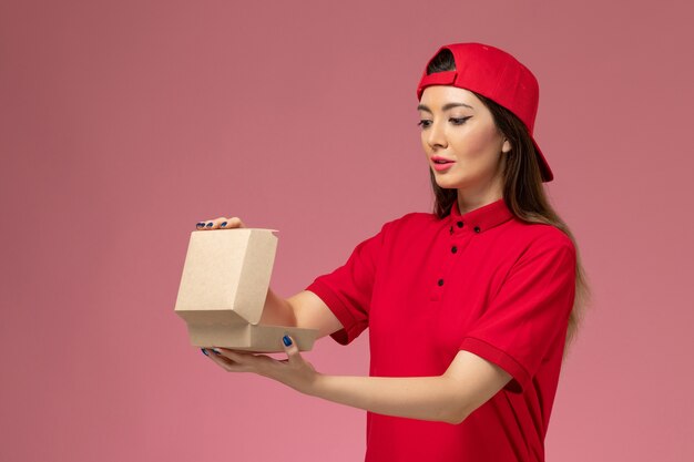 Vooraanzicht jonge vrouwelijke koerier in rood uniform en cape met weinig voedselpakket voor bezorging op haar handen op lichtroze muur