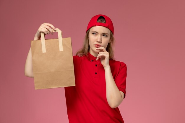 Vooraanzicht jonge vrouwelijke koerier in rood uniform en kaap die voedselpakket houden en aan roze muur denken