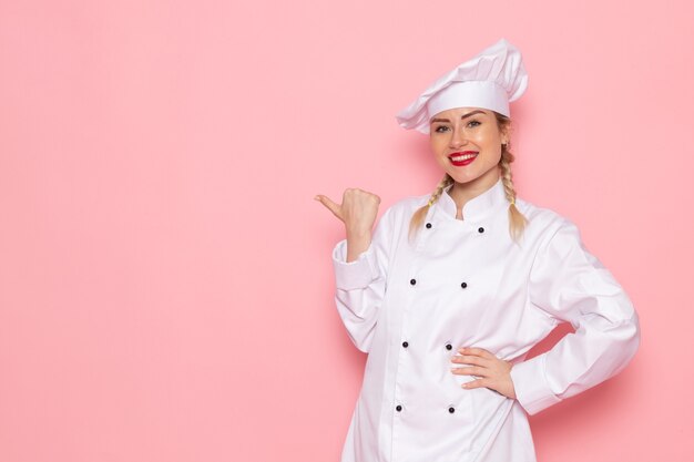 Vooraanzicht jonge vrouwelijke kok in wit kokskostuum die zich enkel bevinden en op de roze ruimte het werkfoto van de kokkeuken stellen stellen