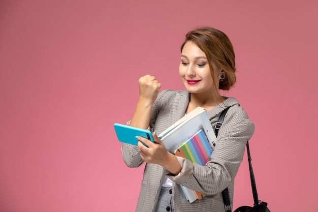 Vooraanzicht jonge vrouwelijke student in grijze jas met voorbeeldenboeken en met behulp van een telefoon met een glimlach op de roze achtergrondlessen universiteit college studie