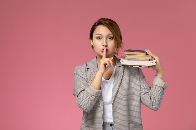 Vooraanzicht jonge vrouwelijke student in grijze jas met voorbeeldenboeken met stilte teken op roze achtergrond lessen universiteit college studie