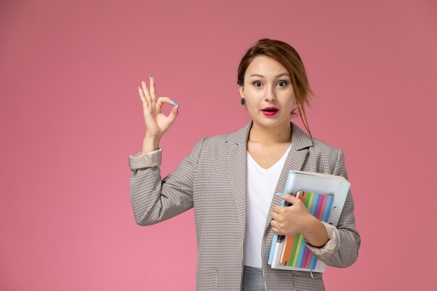 Vooraanzicht jonge vrouwelijke student in grijze jas met voorbeeldenboeken op roze achtergrondlessen hogeschoolstudie