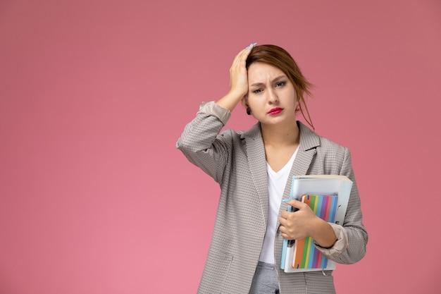 Vooraanzicht jonge vrouwelijke student in grijze jas poseren bedrijf boeken met hoofdpijn op de roze achtergrond lessen hogeschool studie