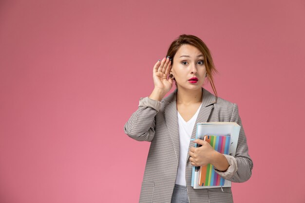 Vooraanzicht jonge vrouwelijke student in grijze jas poseren bedrijf boeken proberen te horen op de roze achtergrond lessen universiteit college studie