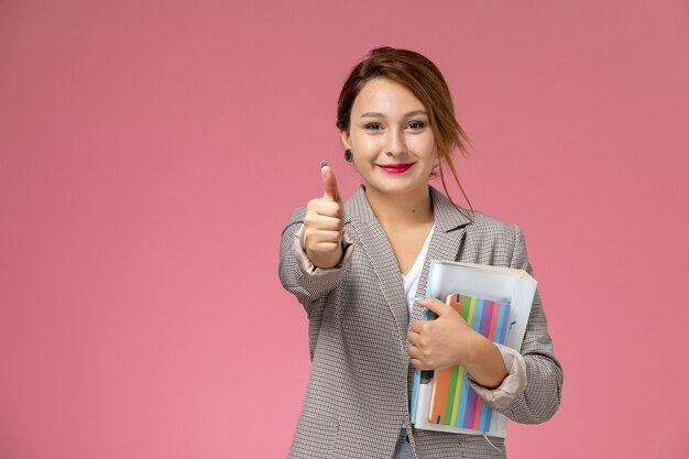 Vooraanzicht jonge vrouwelijke student in grijze vacht poseren bedrijf boeken weergegeven als teken met glimlach op de roze achtergrond lessen hogeschool studie