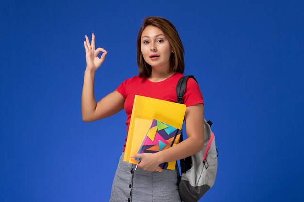 Vooraanzicht jonge vrouwelijke student in rood overhemd die de dossiers en voorbeeldenboek van de rugzakholding op de blauwe achtergrond dragen.