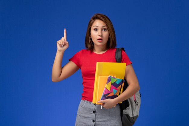 Vooraanzicht jonge vrouwelijke student in rood overhemd die de dossiers van de rugzakholding en voorbeeldenboek dragen die haar vinger op blauw bureau opheffen.