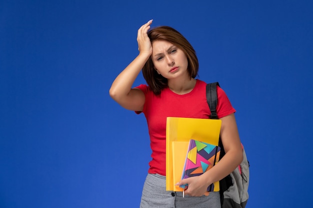 Vooraanzicht jonge vrouwelijke student in rood overhemd die de dossiers van de rugzakholding en voorbeeldenboek dragen met hoofdpijn op blauwe achtergrond.