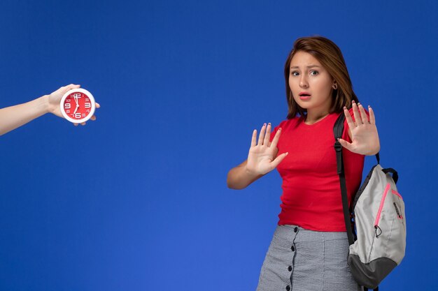 Vooraanzicht jonge vrouwelijke student in rood overhemd die rugzak op de lichtblauwe achtergrond draagt.