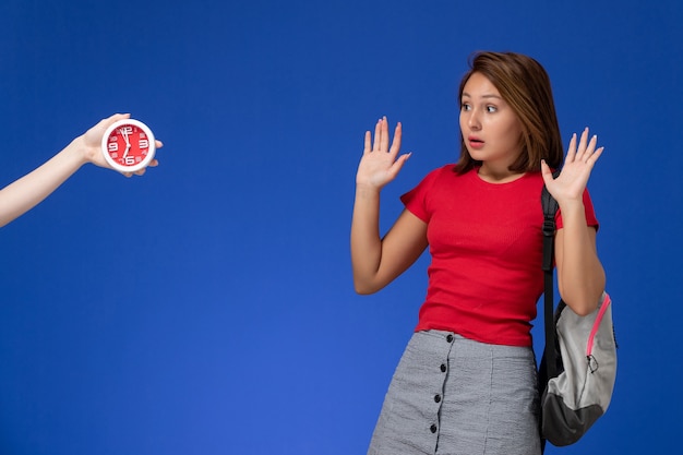 Gratis foto vooraanzicht jonge vrouwelijke student in rood shirt draagt rugzak bang voor klokken op lichtblauwe achtergrond.