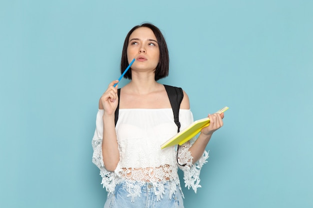 Vooraanzicht jonge vrouwelijke student in wit overhemd, spijkerbroek en zwarte tas denken en voorbeeldenboek op de blauwe ruimte vrouwelijke student universitaire school te houden