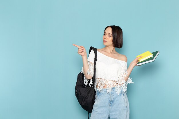 Vooraanzicht jonge vrouwelijke student in wit overhemd, spijkerbroek en zwarte tas met voorbeeldenboeken op de blauwe ruimtestudent