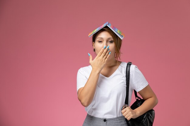 Vooraanzicht jonge vrouwelijke student in wit t-shirt en grijze broek met voorbeeldenboek op haar hoofd op de roze achtergrondlessen hogeschool