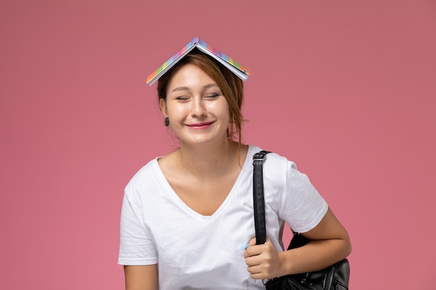 Vooraanzicht jonge vrouwelijke student in wit t-shirt glimlachend bedrijf tas op roze achtergrond les universiteit college studieboek