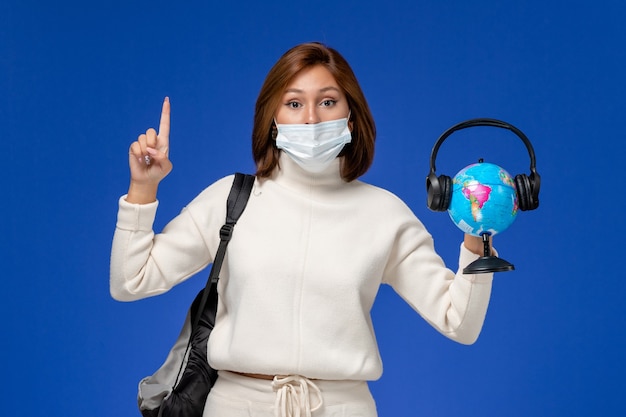 Vooraanzicht jonge vrouwelijke student in witte trui dragen masker en rugzak met globe met koptelefoon op de blauwe muur